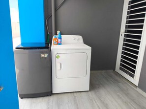 Ein kleiner Waschraum mit einer grauen Waschmaschine und einem weißen Trockner, einer blauen Wand und einem Fenster mit Jalousien.