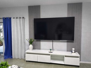 Un salon moderne avec un téléviseur à écran plat fixé au mur, un meuble de rangement blanc avec des tiroirs et un rideau bleu et blanc.