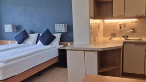 A compact hotel room with a bed, bedside table, and a kitchenette area including a sink and cabinets.