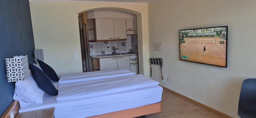 Habitación de hotel con cama, televisor montado en la pared, kitchenette y puerta que da a otra habitación.
