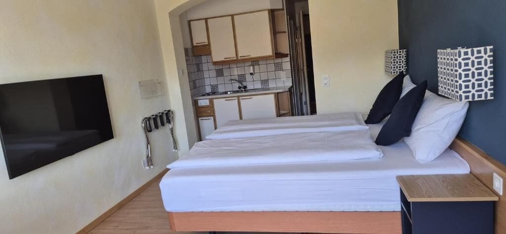 Un dormitorio con cama, televisor montado en la pared, una kitchenette pequeña y un baño visible a través de una puerta abierta.