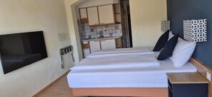 Un dormitorio con cama, televisor montado en la pared, una kitchenette pequeña y un baño visible a través de una puerta abierta.