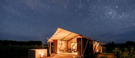 Estructura tipo tienda de campaña con cielo estrellado visible a través de sus aberturas, un pequeño hogar para fogata y un sendero que lleva a la entrada.