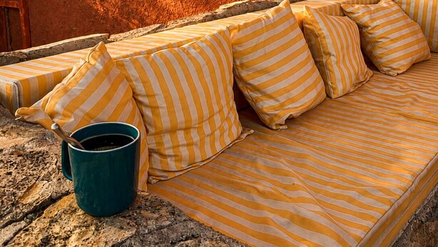 A stone bench with a striped cushion and a green bucket, positioned in front of a building with a textured orange wall and a window with green shutters.