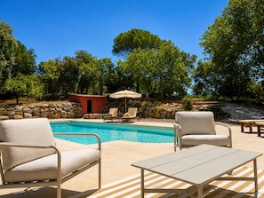 A swimming pool with lounge chairs and a small red building, surrounded by trees.