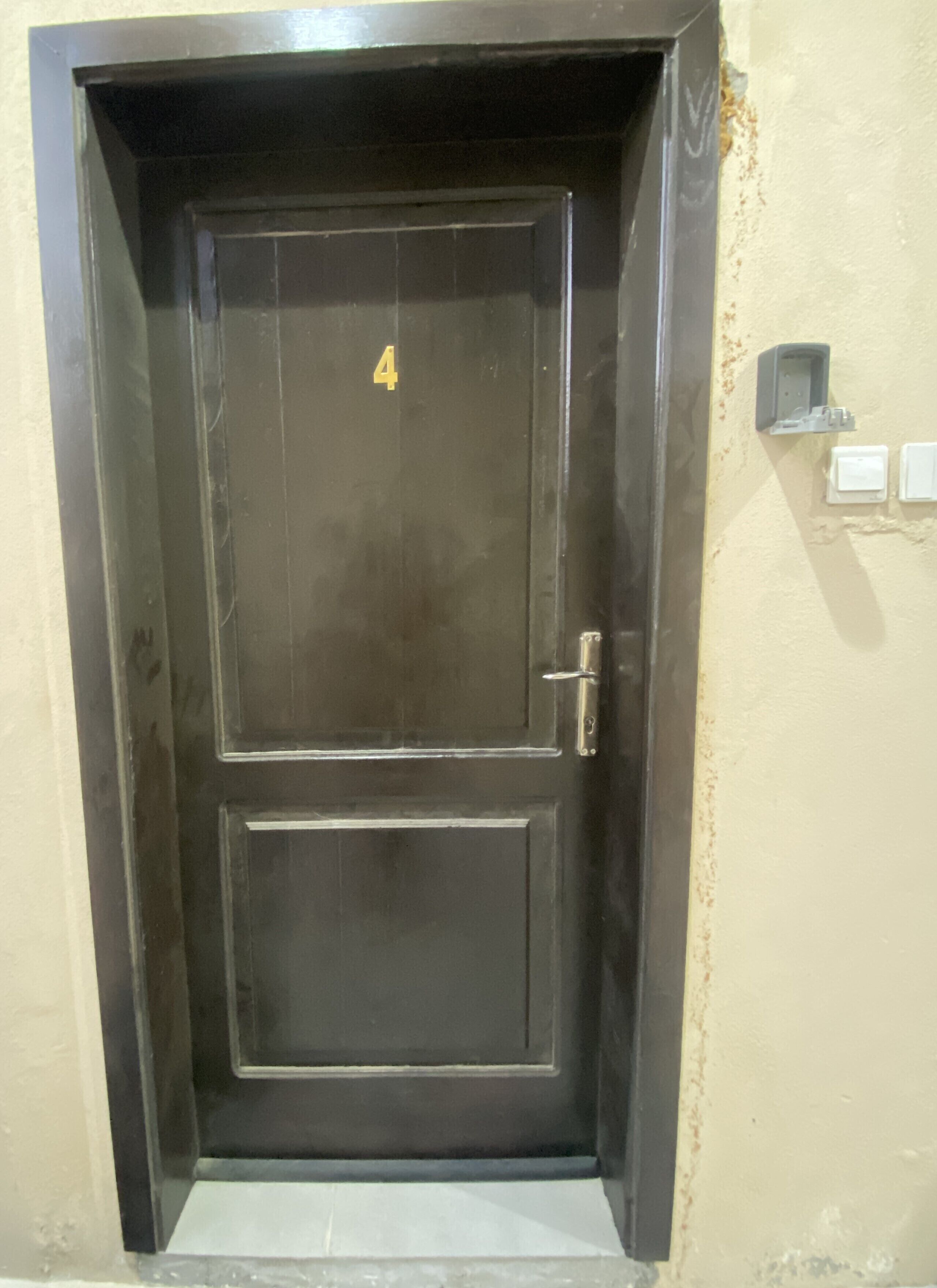 Una puerta de madera oscura con el número 4, un picaporte y una pequeña caja eléctrica en la pared a la derecha.
