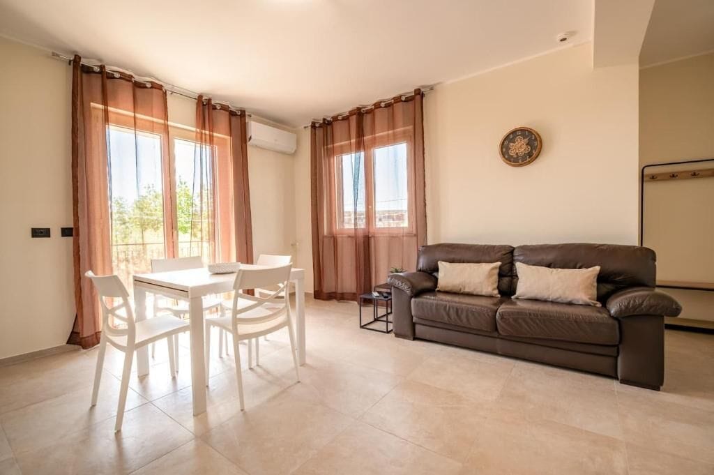 Ein Wohnzimmer mit braunem Ledersofa, einem Esstisch mit Stühlen und großen Fenstern mit Vorhängen.