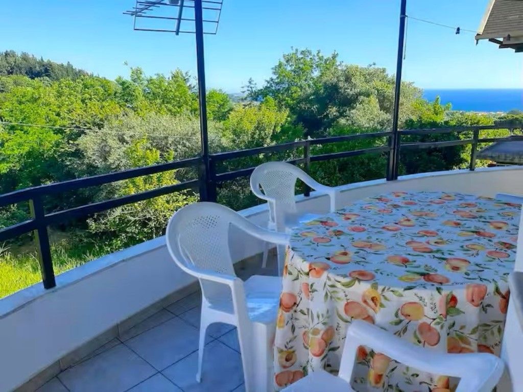 A balcony with a table and chairs, overlooking a forested area and the sea.