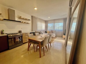 A modern kitchen with a stove, dining table, and chairs. There is a sofa and a window with curtains. - Casa Amber - A designer retreat in the heart of authentic Mexico (Puerto Morelos)