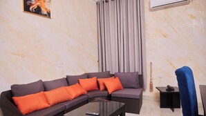 Una sala de estar con un sofá modular, una mesa de centro de vidrio, un control remoto, un sillón azul y un equipo de aire acondicionado instalado en la pared.