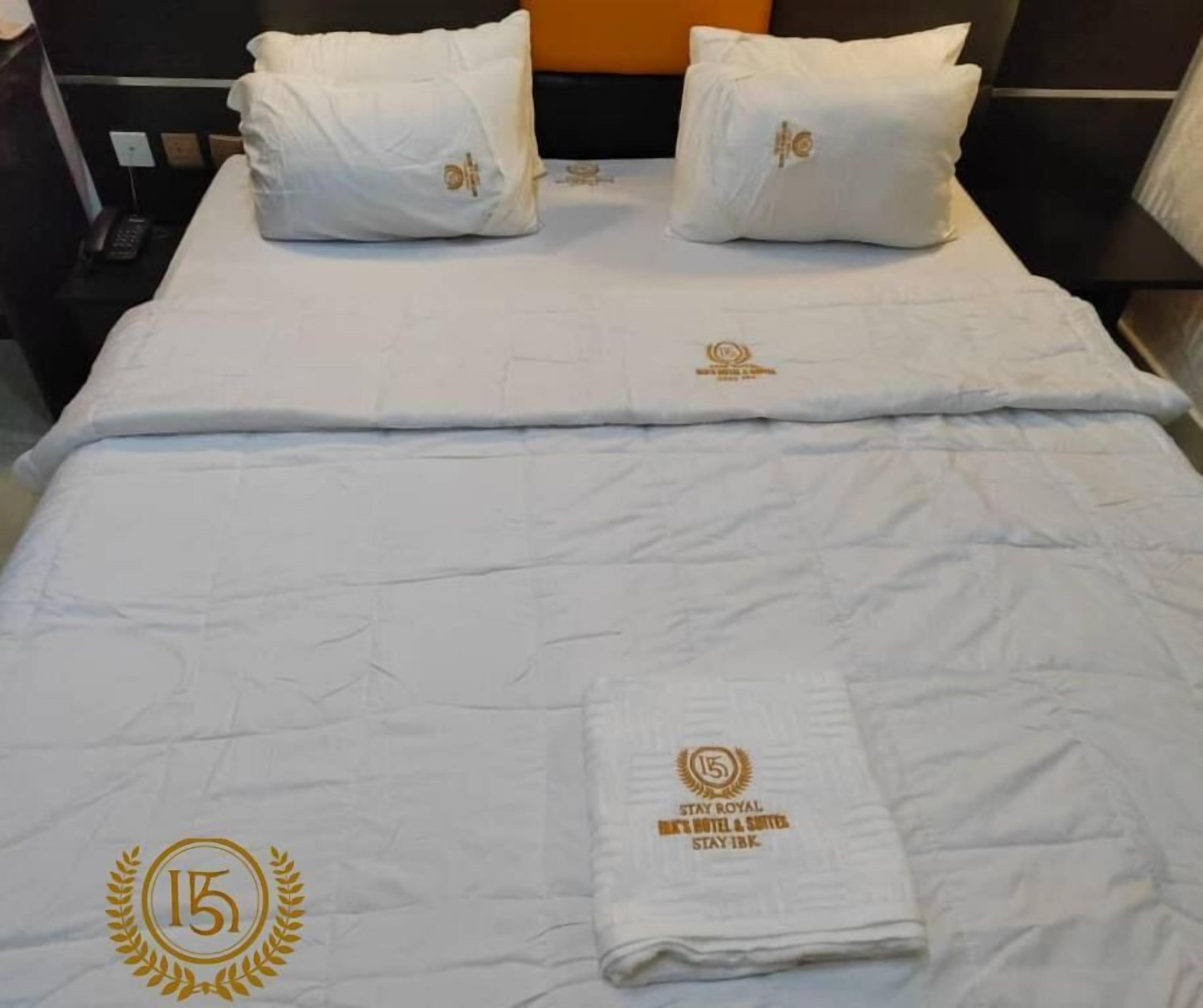 Quarto de hotel com uma cama bem arrumada, uma toalha dobrada com o logotipo do hotel e uma mesa de cabeceira com um telefone.