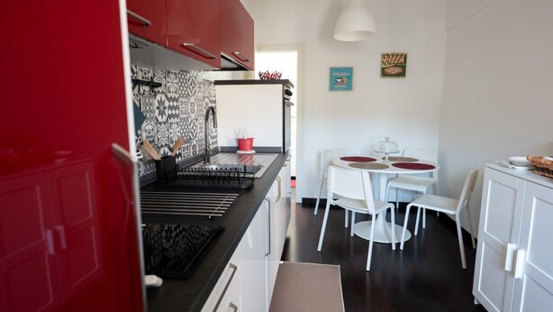 A compact kitchen with a red cabinet, a black countertop, and a white dining area with two chairs and a small table. - GuestHost - Casa Arezzo al Duomo (Modica)