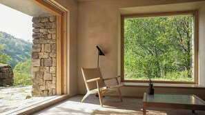A room with a stone wall, a large window with a view of trees, a wicker chair, and a floor lamp. - Designed and renovated villa with mountain views (Selaya)