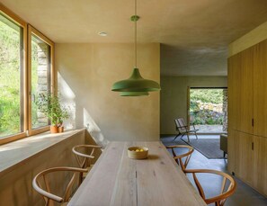 A modern dining area with a wooden table, chairs, a hanging green pendant light, and a view of greenery outside. - Designed and renovated villa with mountain views (Selaya)