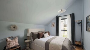 Un dormitorio con cama, una silla, una mesita de noche, una lámpara, un cuadro en la pared y una ventana con cortinas.
