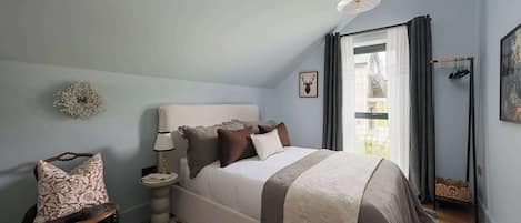 A bedroom with a bed, a chair, a bedside table, a lamp, a picture on the wall, and a window with curtains.