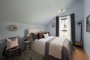 Ein Schlafzimmer mit Bett, Sessel, Nachttisch, Lampe, einem Bild an der Wand und einem Fenster mit Vorhängen.