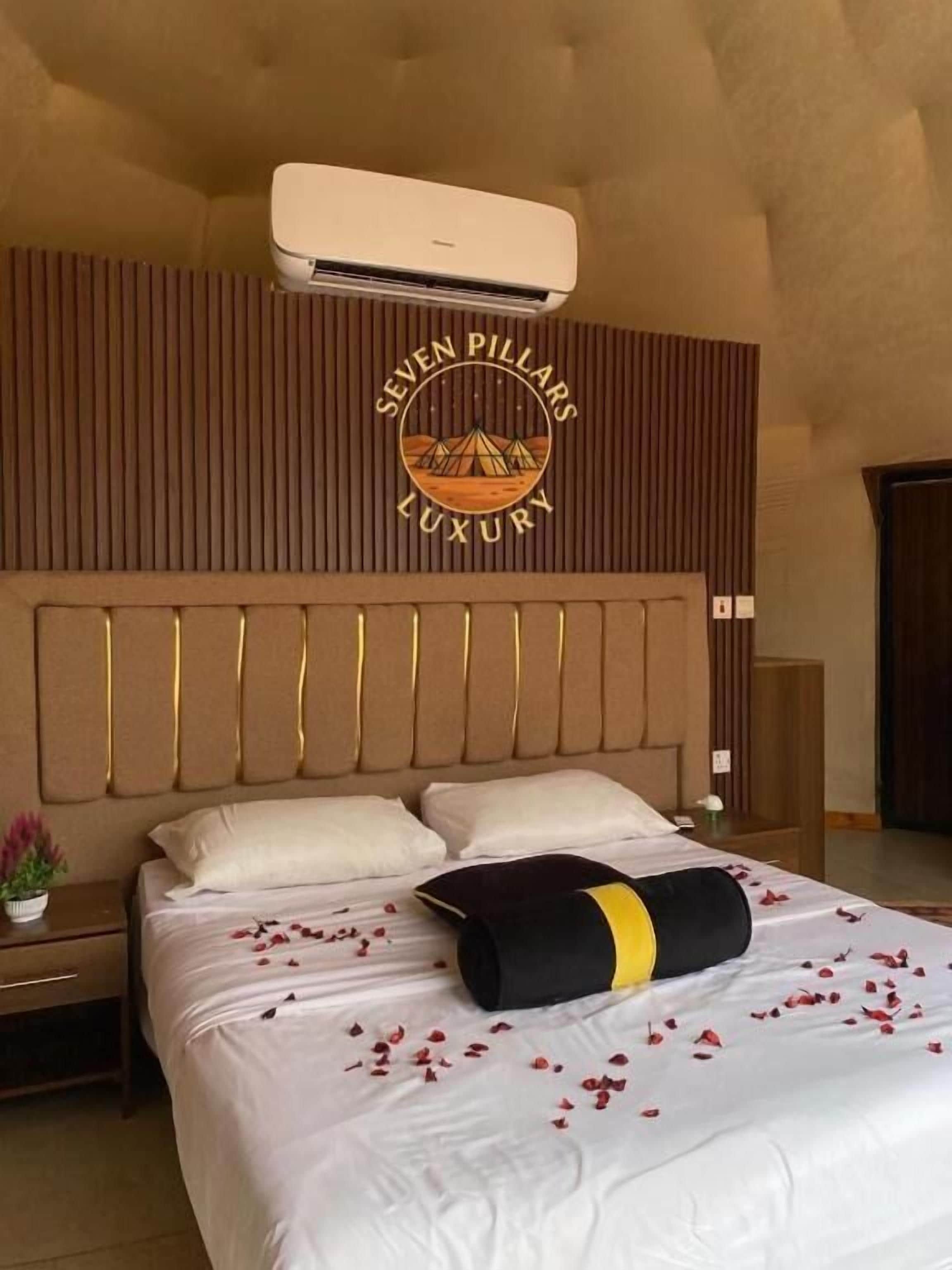 A hotel room with a large bed, a headboard with vertical slats, an air conditioning unit, and a logo on the wall.