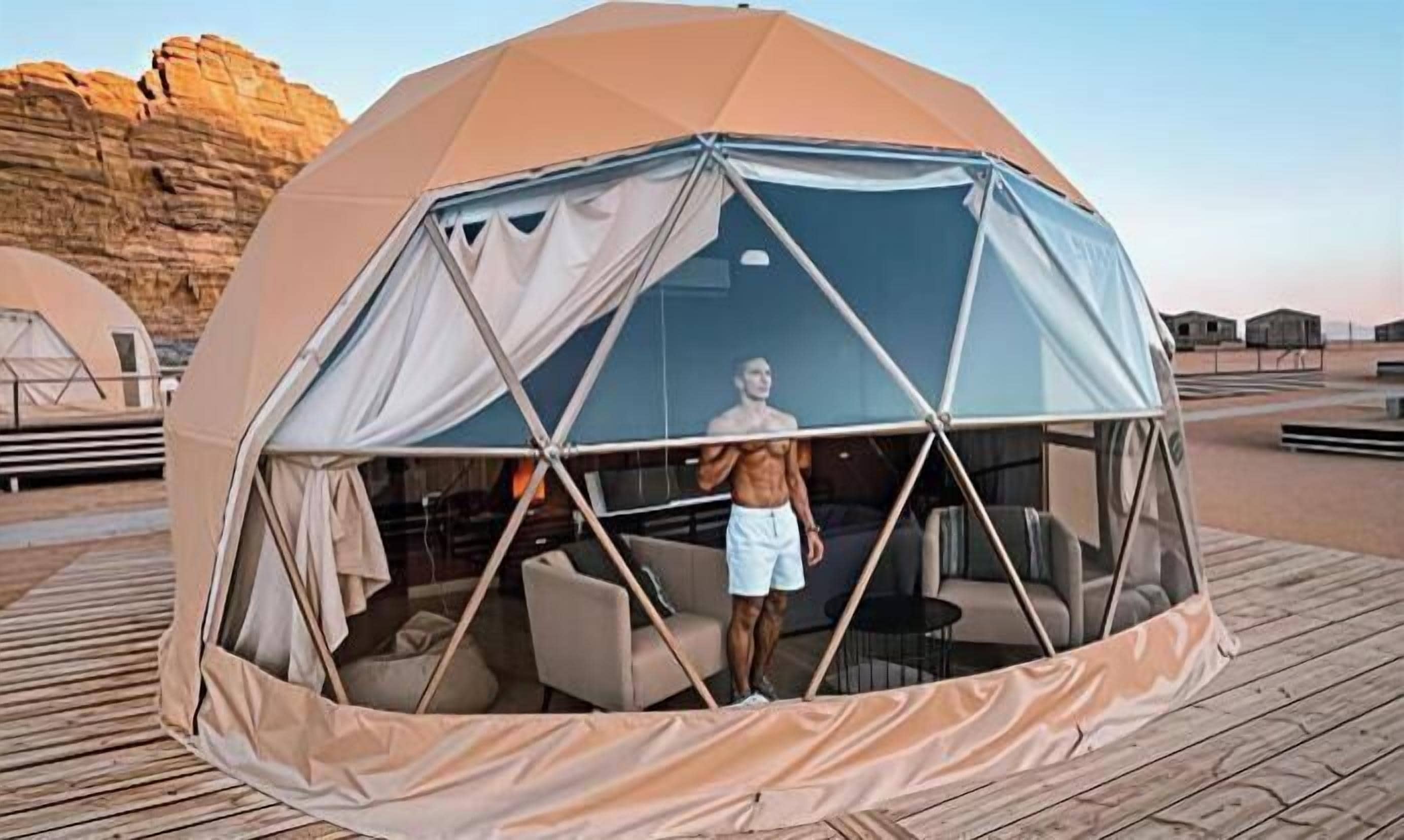 Una tienda de campaña con forma de cúpula, una persona de pie en su interior, una terraza de madera y un paisaje rocoso al fondo.