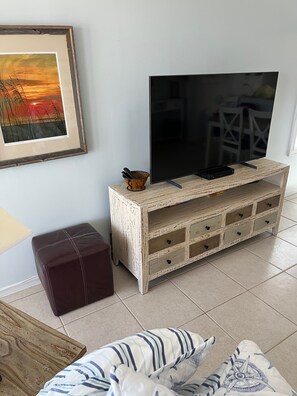 Ein Wohnzimmer mit Fernseher auf einer Holzkommode, einem gerahmten Bild an der Wand und einem braunen Ottomanen.
