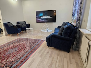 Un salón con un televisor de pantalla plana montado en la pared, dos sillones azules, una mesa de centro redonda y una alfombra estampada.