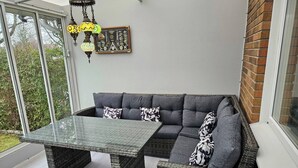Een modern buitenzitgedeelte met een tafel met glazen blad en rieten meubilair, een decoratieve wanddecoratie en uitzicht op groen.