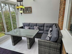 Ein moderner Außenbereich zum Sitzen mit einem Glastisch, Korbmöbeln, einer Wanddekoration und Blick auf viel Grün.