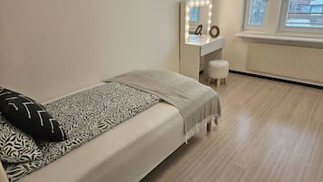 A bedroom with a bed, a vanity with a mirror and lights, a small stool, and a window with blinds.