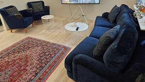 Een woonkamer met een televisie, twee blauwe fauteuils, een blauwe bank, een tapijt met een patroon en een kleine witte tafel met een afstandsbediening.