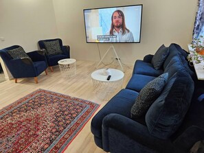Ein Wohnzimmer mit Fernseher, zwei blaue Sessel, ein blaues Sofa, einem gemusterten Teppich und einem kleinen weißen Tisch mit einer Fernbedienung.