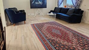 Een woonkamer met een flatscreen televisie, een blauwe bank, een kamerkleed met een patroon en een raam met bloemen gordijnen.