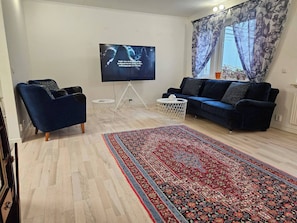 Ein Wohnzimmer mit einem Flachbildfernseher, einem blauen Sofa, einem gemusterten Teppich und einem Fenster mit floralen Vorhängen.