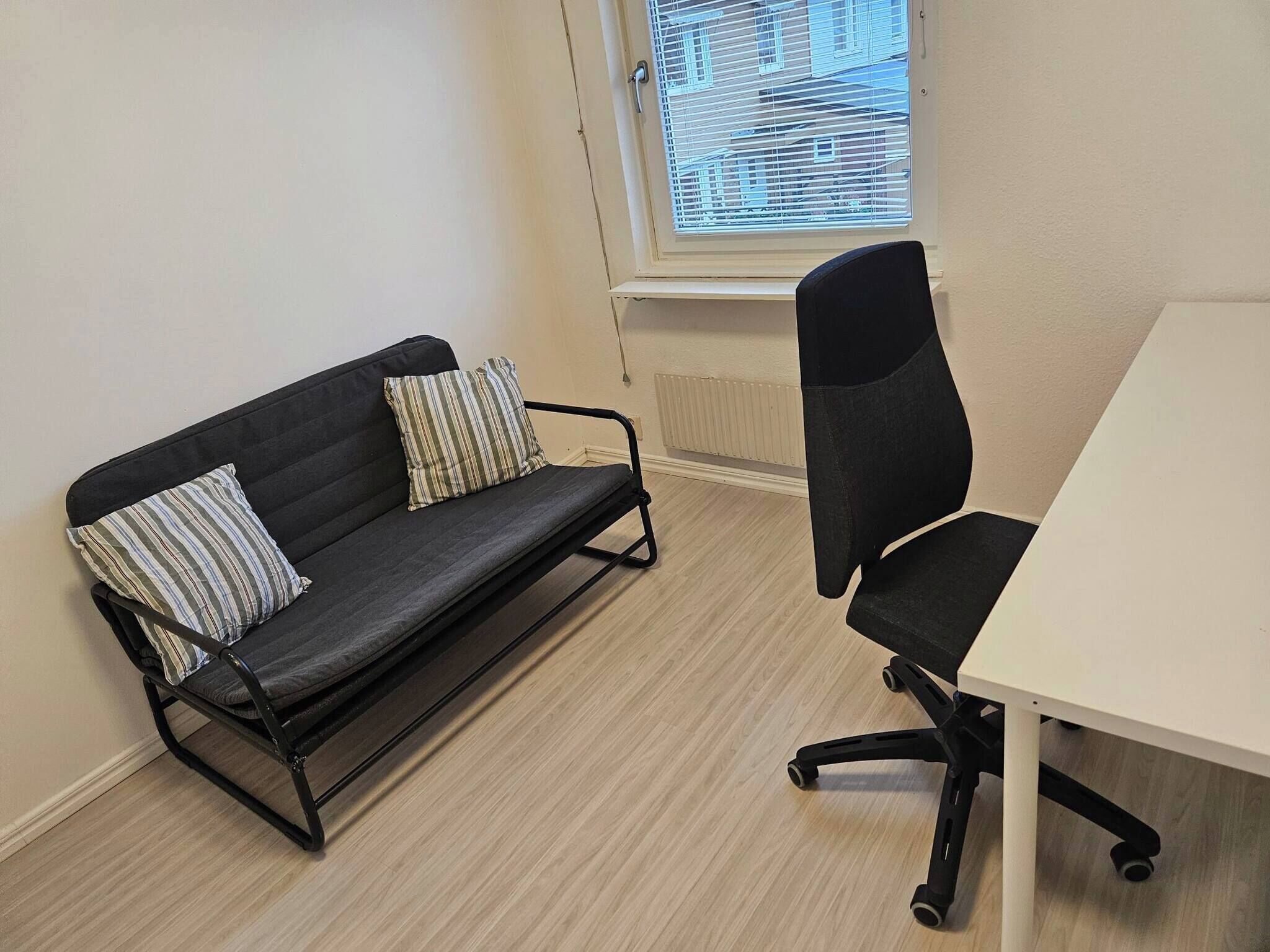 Una habitación con un sofá negro, dos almohadas a rayas, una silla de oficina negra y una mesa blanca.