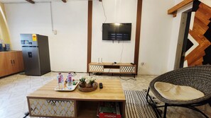 A living room with a television mounted on the wall, a wooden coffee table with a board game, a wicker chair, and a refrigerator. - 1-BR Quiet  Private Stay Peaceful Atmosphere (Accra)
