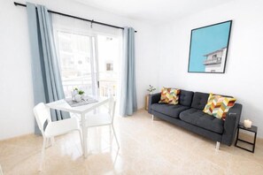 A modern living room with a dining area, a grey sofa with colorful cushions, a framed picture on the wall, and a view of the outside through a large window. - Edificio Don Diego (Los Cristianos)