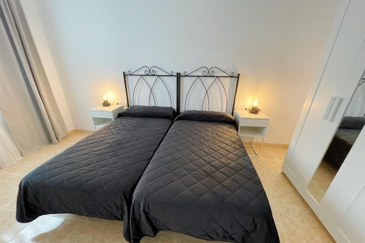 Una cama doble con edredón acolchado negro, dos mesitas de noche con lámparas y un armario con espejo.