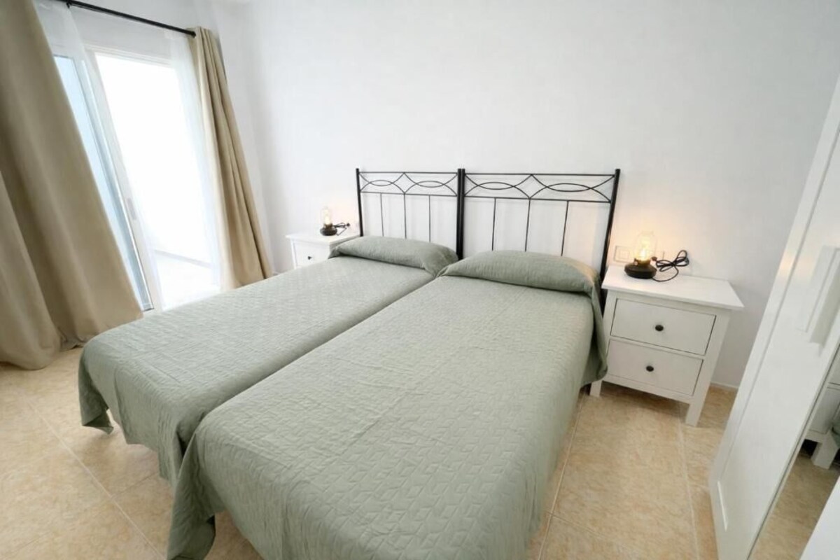 Una cama doble con estructura metálica, dos mesitas de noche con lámparas y una ventana con cortinas.