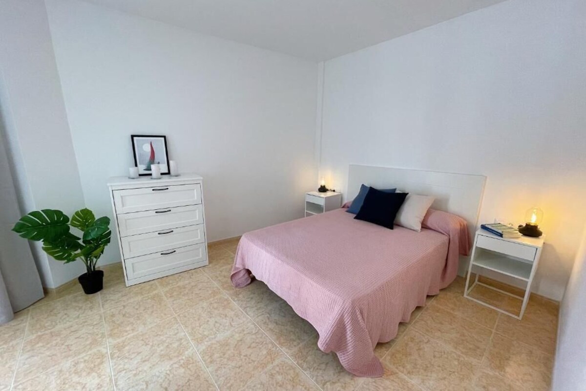 Un dormitorio con cama, mesitas de noche, cómoda, planta en maceta y mesita con lámpara.