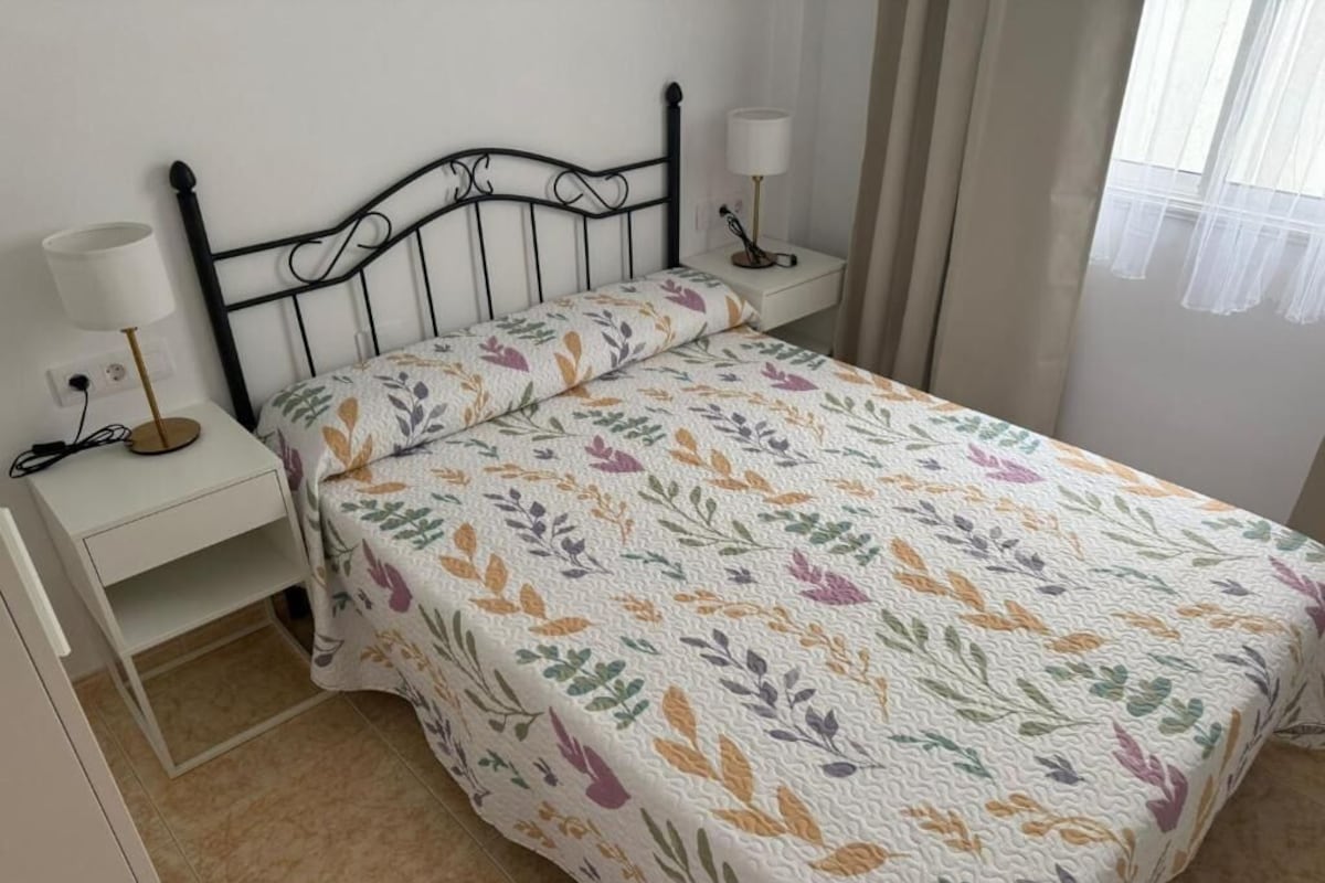 Un dormitorio con cama, mesita de noche, lámpara y ventana con cortinas.
