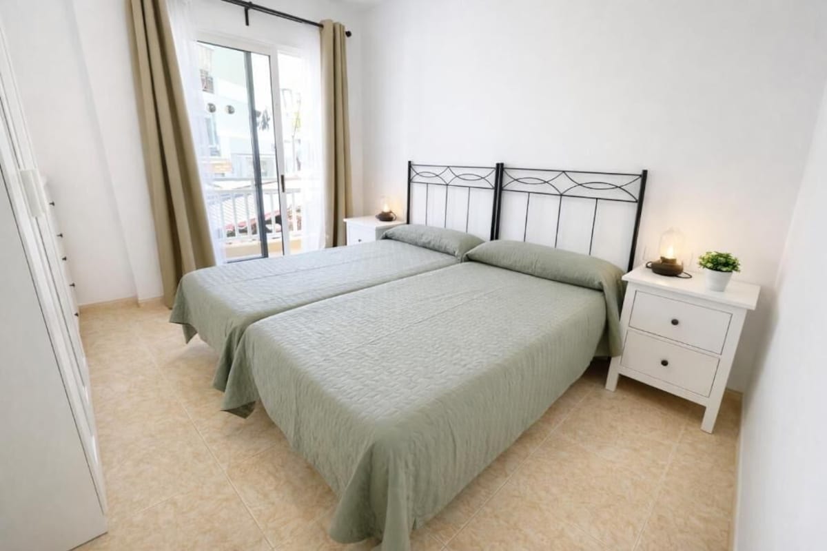 Un dormitorio con dos camas, una mesita de noche blanca y una ventana con cortinas.