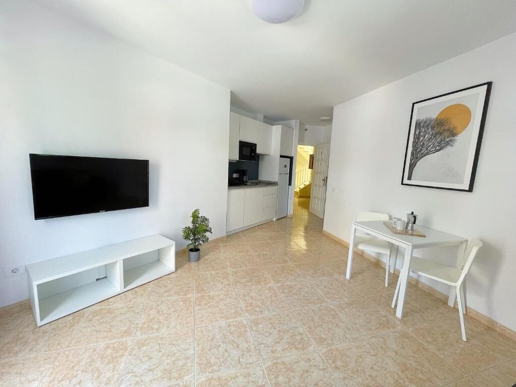 A modern living room with a flat-screen TV, a dining area with a table and chairs, and a kitchen in the background.