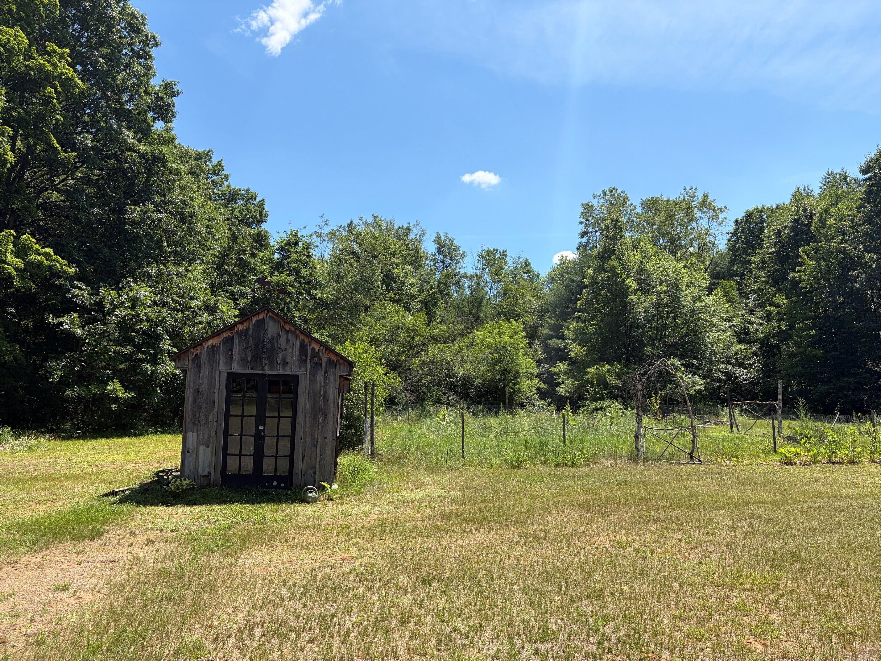 A rustic wooden shed in a grassy field with trees in the background.