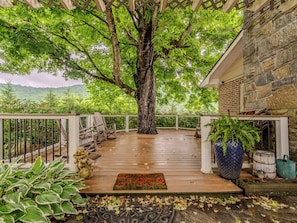 Una terraza de madera con barandal blanco, un árbol grande y un edificio de piedra al fondo.