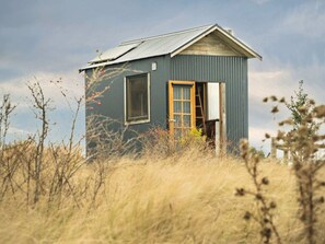 A small, single-story building with a gabled roof, a door, and a window, surrounded by tall grass and sparse vegetation.