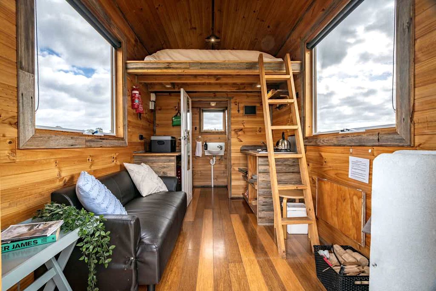 A cozy cabin interior with a wooden floor, a sofa, a ladder, and a small kitchenette.