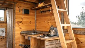 A wooden cabin interior with a kitchen area, including a sink, stove, kettle, and a ladder.