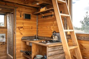 A wooden cabin interior with a kitchen area, including a sink, stove, kettle, and a ladder.
