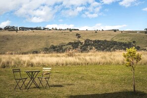 An outdoor seating area with two chairs and a table, overlooking a grassy field and distant trees under a partly cloudy sky.