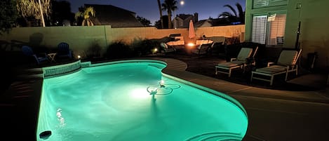 A swimming pool at night with a green light, surrounded by lounge chairs and a patio area.