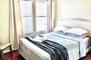 Ein ordentlich bezogenes Bett mit Kopfteil, Nachttisch und einem Fenster mit Jalousien.